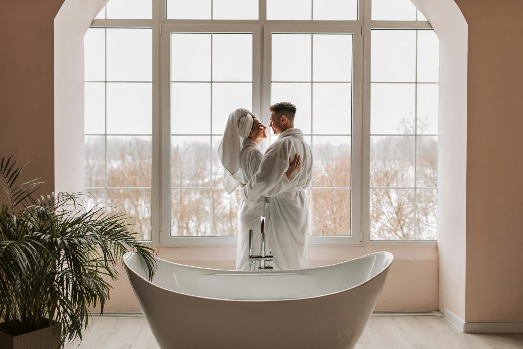 Couple in bathrobes standing together by a freestanding tub, enjoying a romantic spa moment by large windows